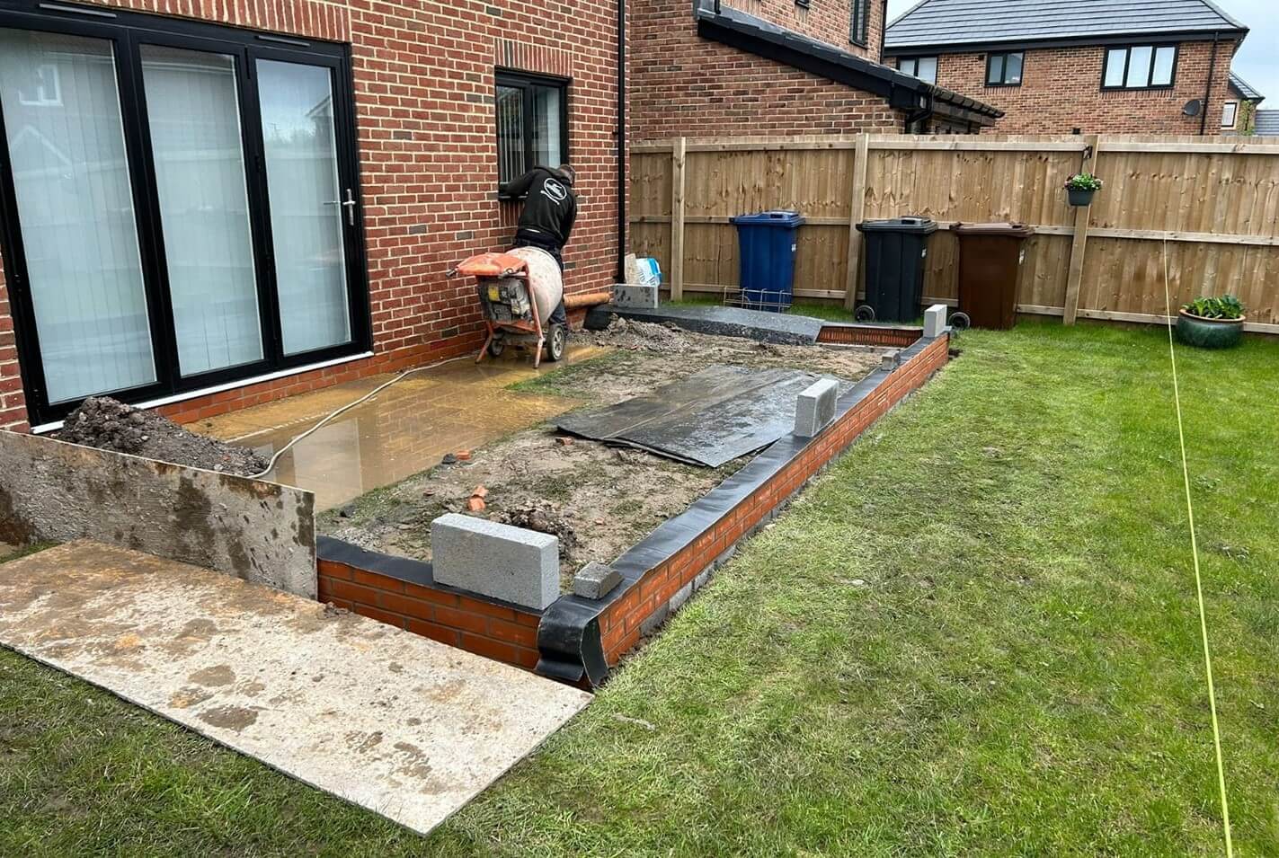 A brick patio with a wooden bench and a fence, with a brick wall being built in the garden for extension.