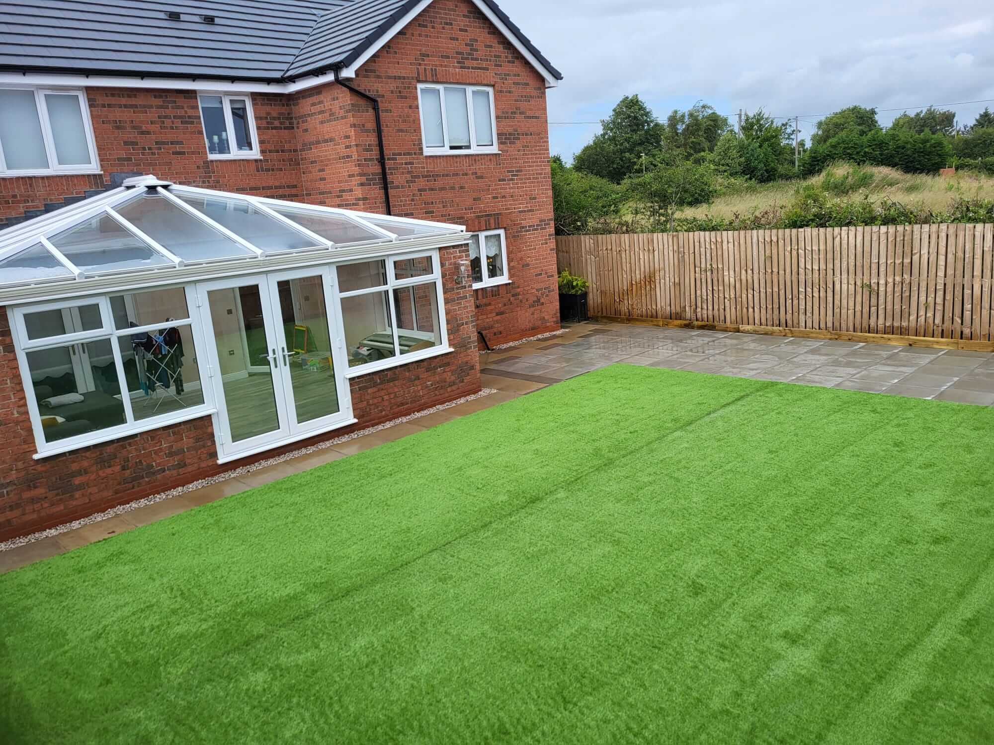 Backyard of a brick house featuring a modern glass conservatory extension and freshly landscaped area with lush green artificial grass and a paved patio