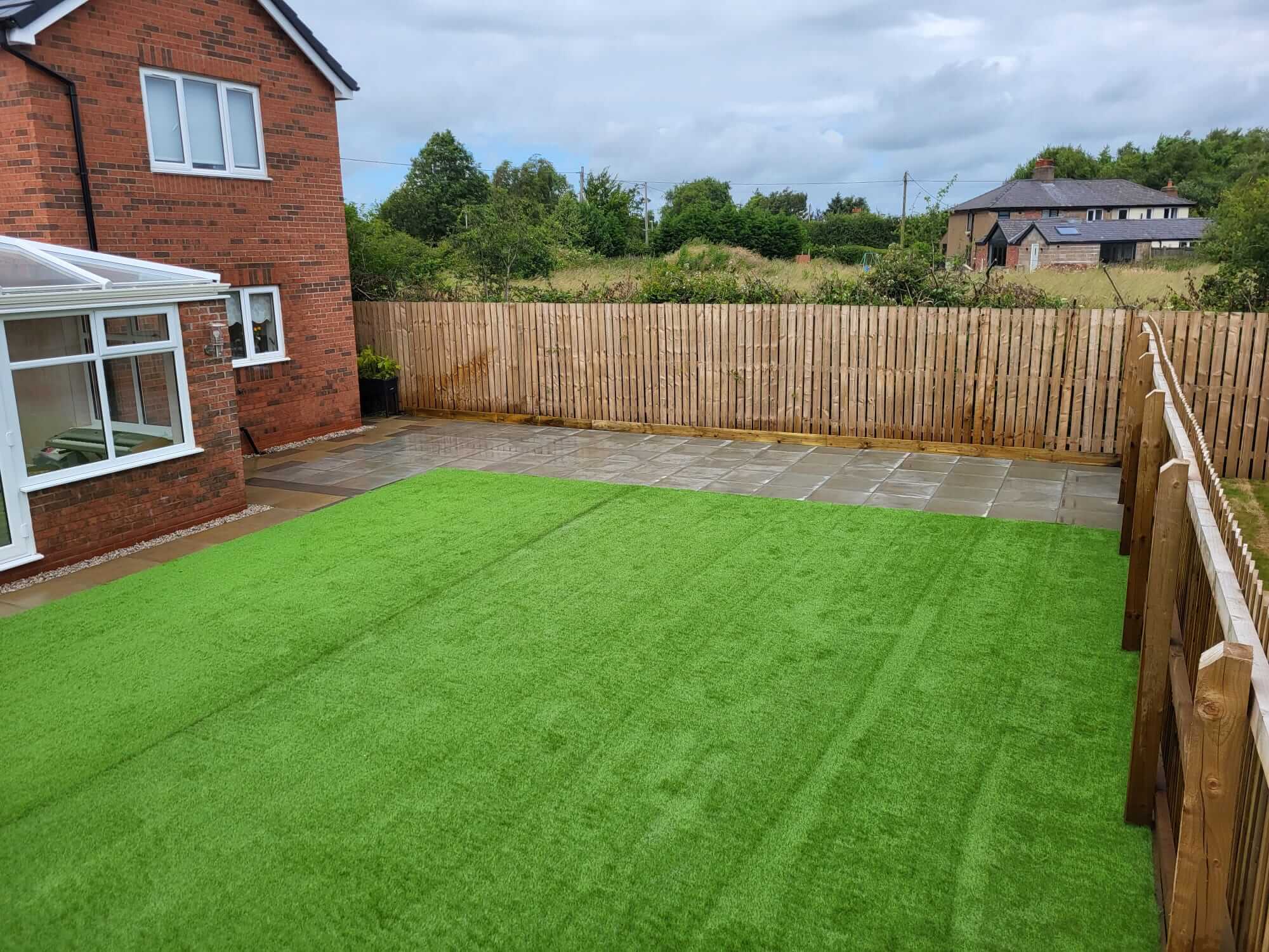Side view of a brick house showcasing a new glass conservatory extension and a freshly landscaped garden with artificial grass and a paved patio area, enclosed by a wooden fence.