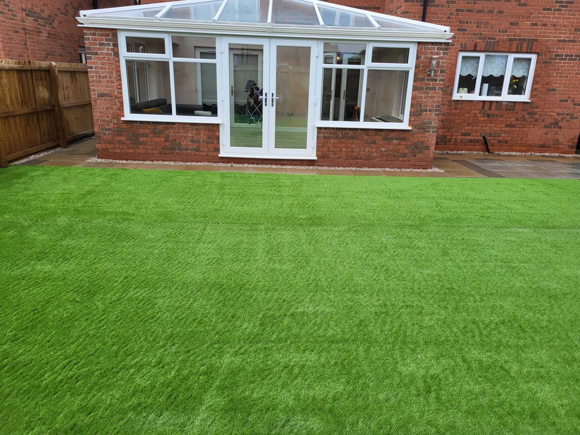 A backyard featuring a bright green artificial lawn leading up to a red brick house with a white conservatory.